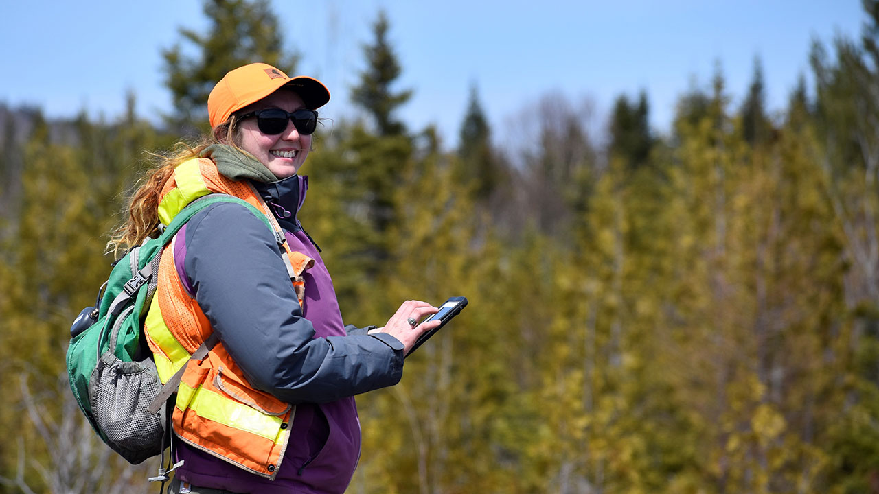 Tetra Tech employee with long hair in orange hat and vest types on a mobile device during a wind project field survey
