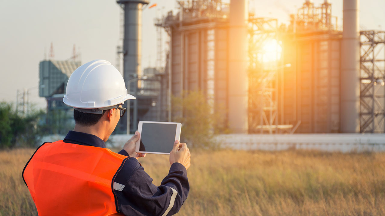 Engineer in hard hat and orange vest with iPad in front of power plant