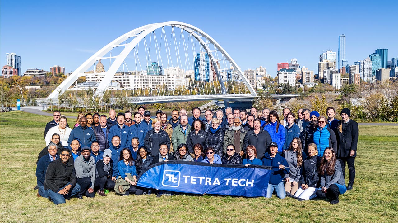 A large group of employees holding a Tetra Tech banner at a park with a city skyline and bridge in the background