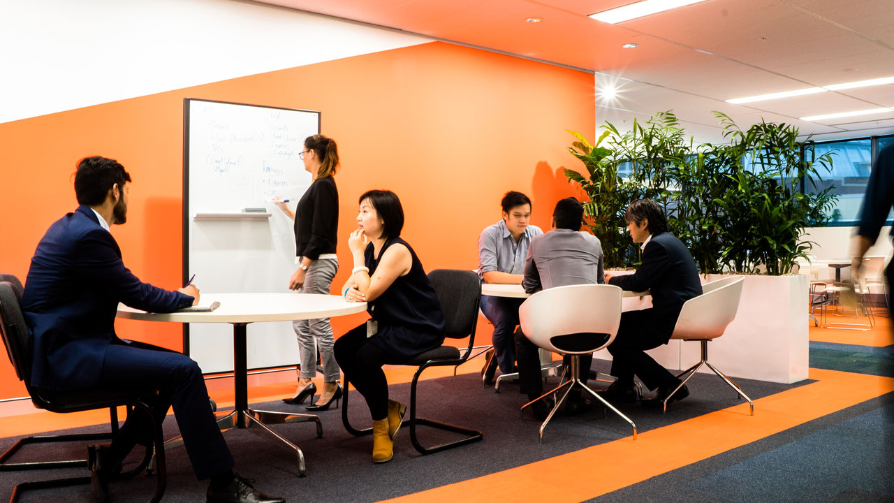 A woman writes on a white board, and people sit in groups around tables in a casual collaborative workspace with orange walls
