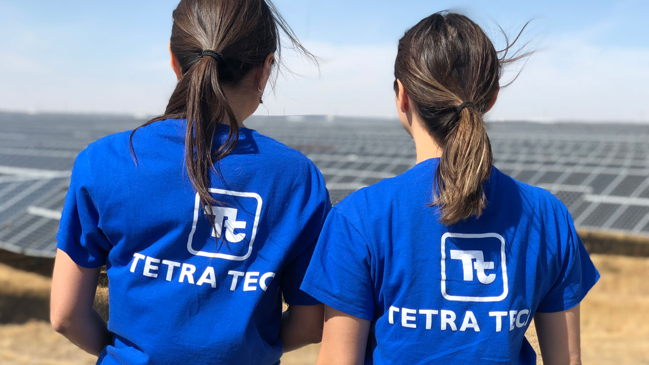 Two women with their backs to the camera wearing blue Tetra Tech shirts look out over a solar farm in Kazakhstan