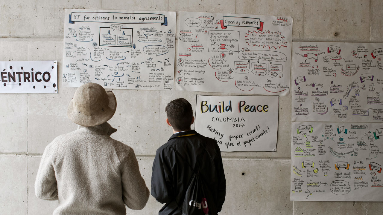 Two men look at a wall covered in posters and graphical drawings, one including the text “Build Peace Colombia”