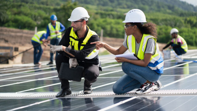 Two people wearing yellow vests and hard hats crouching on solar panels
