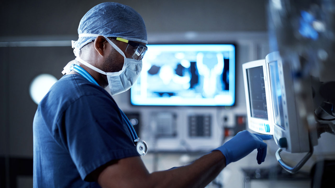 A doctor in surgical scrubs and protective equipment checks computer displays