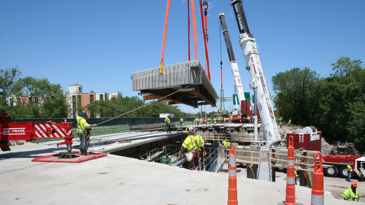 A large pre-cast modular panel with two steel plate girders and reinforced concrete deck, being erected with heavy lift equipment