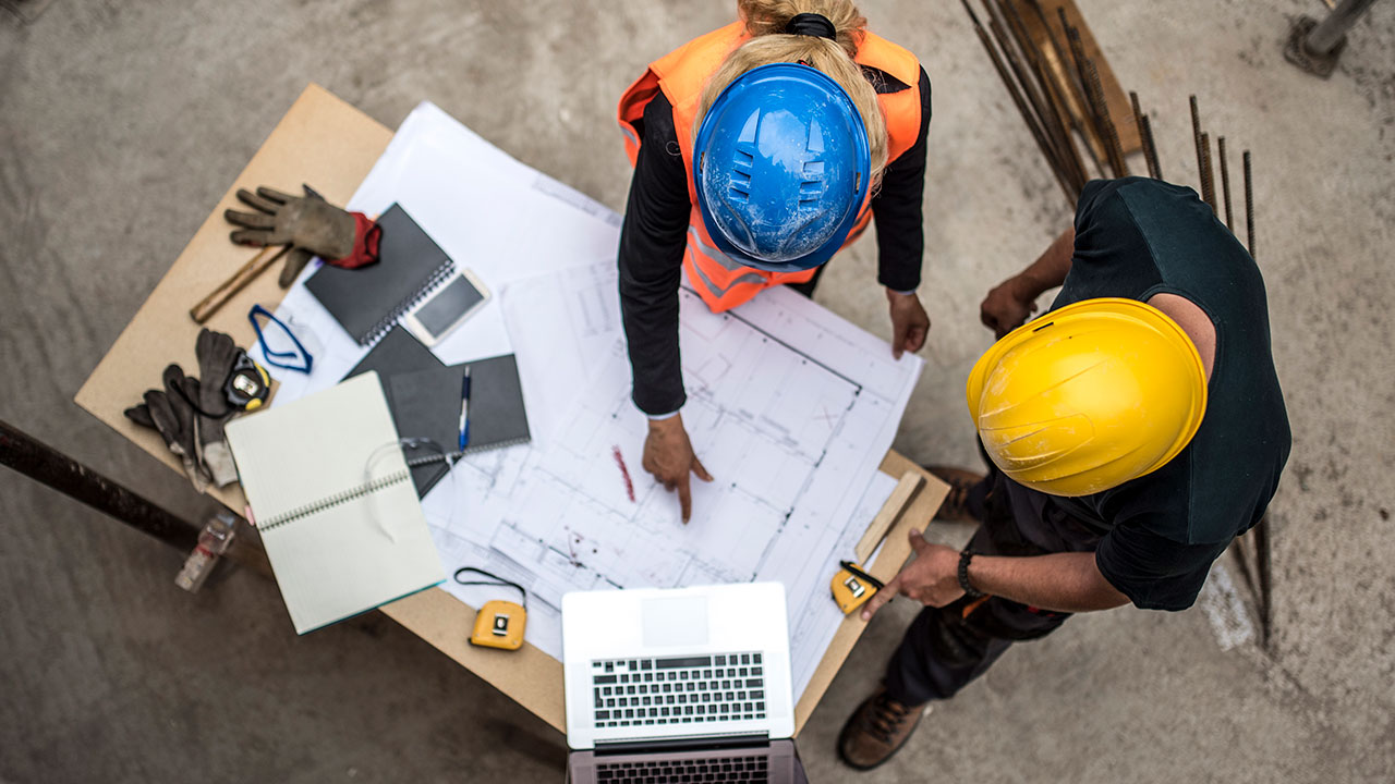 Overhead view of two people with hard hats standing around a table and looking at drawing plans