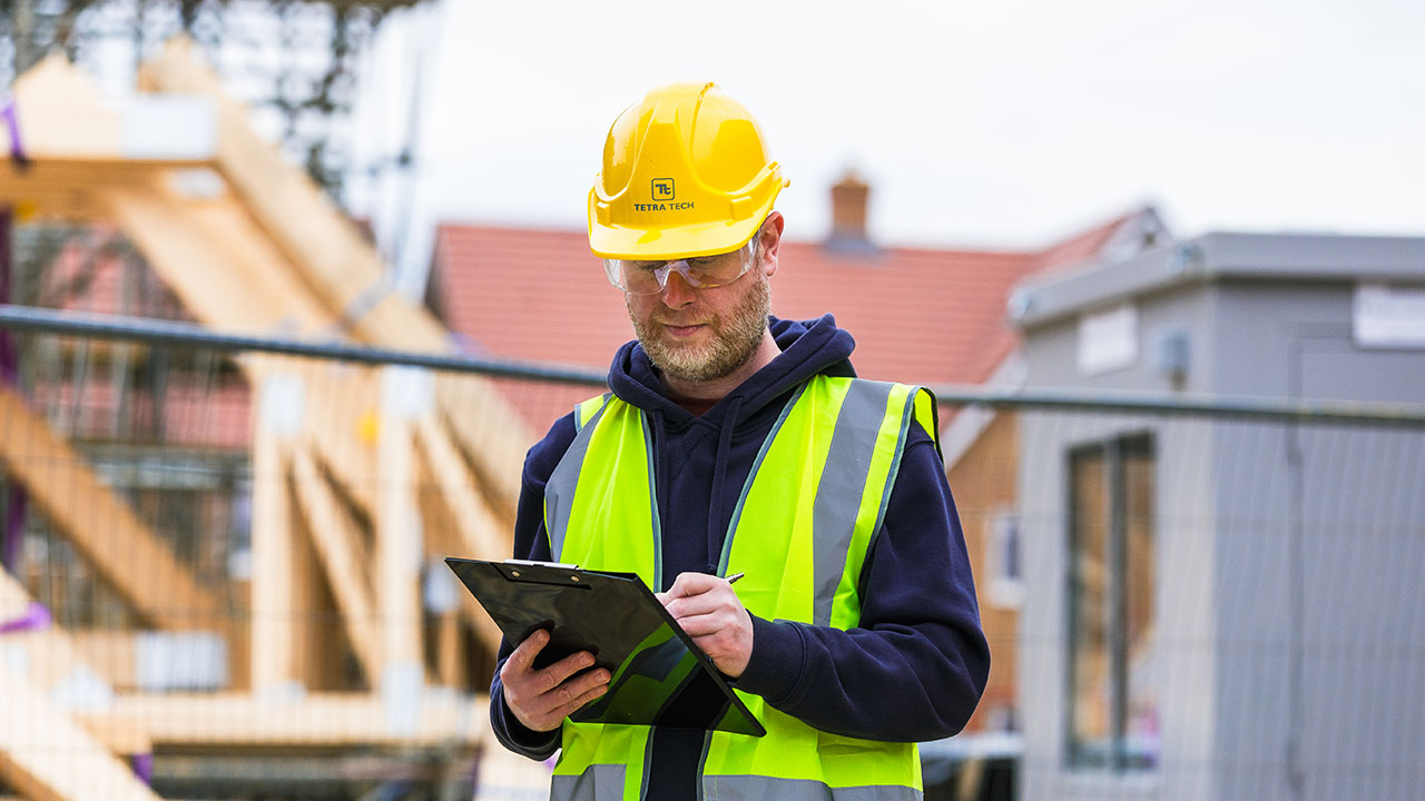 Worker wearing a hard hat, goggles, and safety vest examining documents on a clipboard with the job site in the background