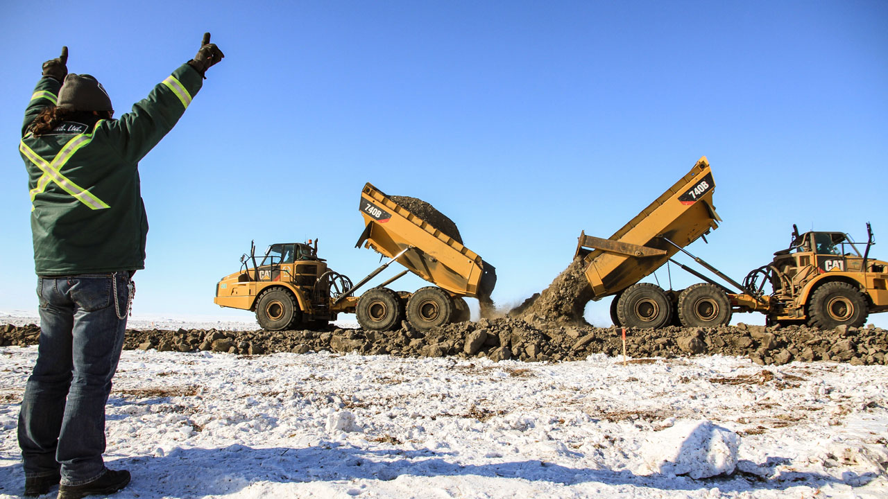 A person in winter gear in the foreground of a snowy field raises both arms in the air while two dump trucks offload dirt for an all-weather road