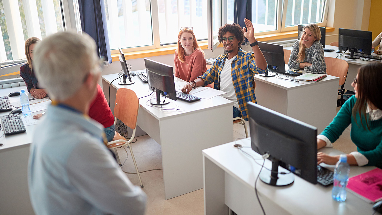 Student at a computer in a classroom raises hand to ask a question to the instructor