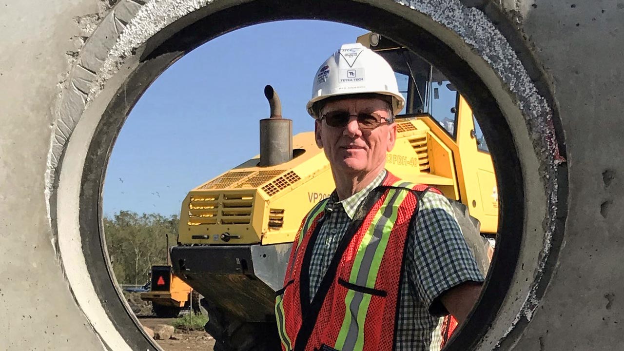 Worker posing for a photo wearing a hard hat, glasses, and safety vest with machinery in the background