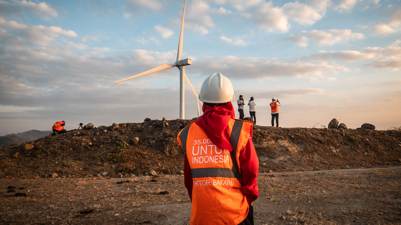 A female wind power engineer stands looking at a windmill in the distance in Indonesia