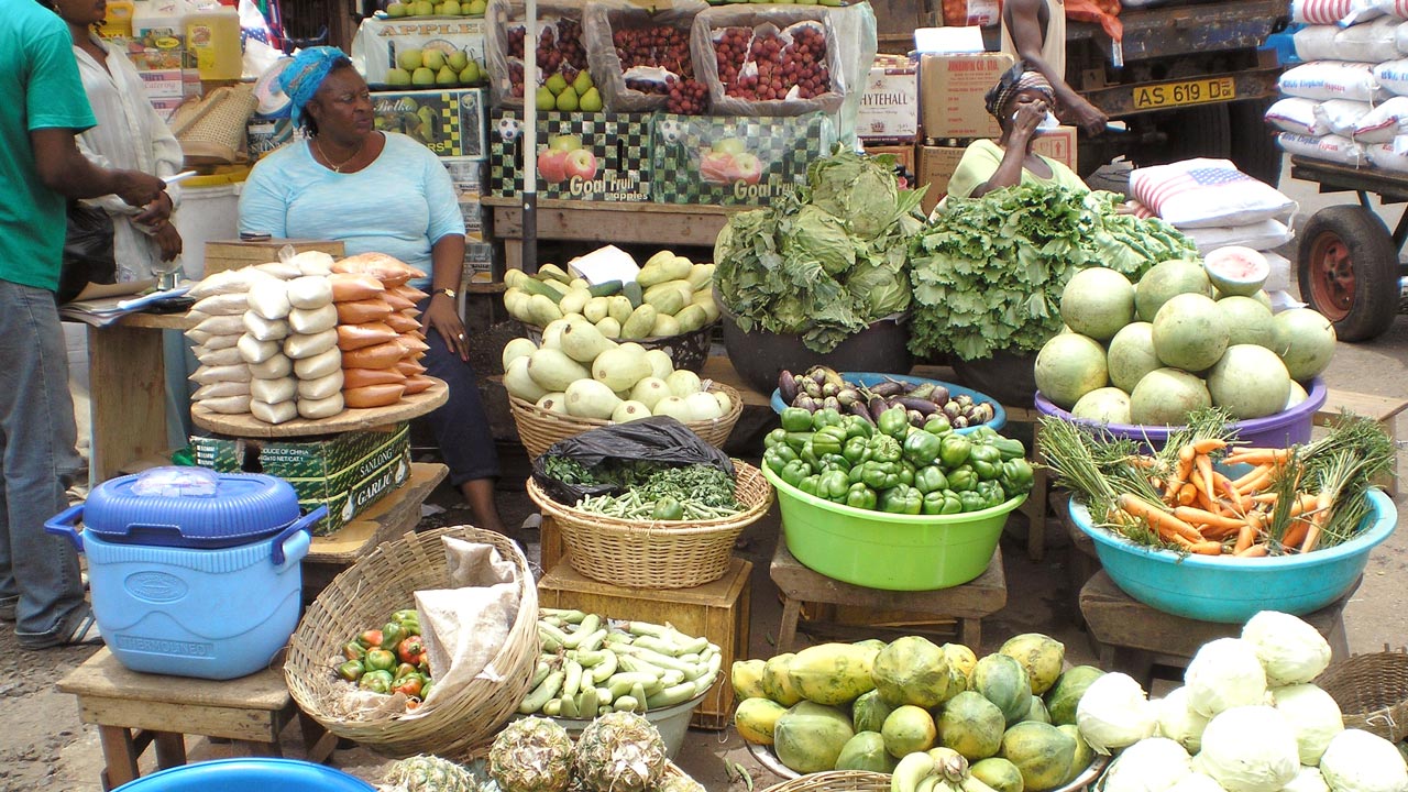 A woman sits behind buckets of various vegetables at her stall at a market in Ghana