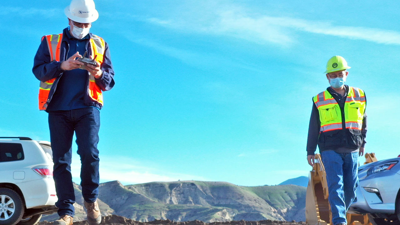 Two people wearing safety vests, hard hats, and masks, walk between parked cars