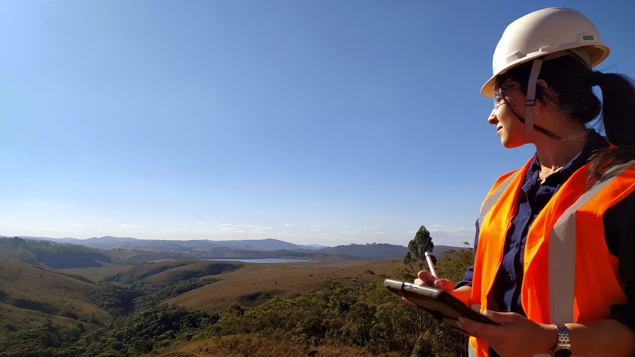 A woman wearing a safety vest and hard hat holding a clipboard stands on a hill looking out at the terrain