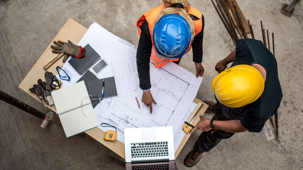 Overhead view of two people with hard hats standing around a table and looking at drawing plans