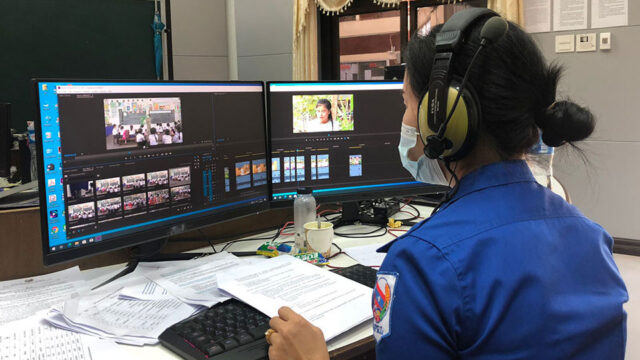 A woman wearing headphones sits at a desk with her back to the camera, working on a dual screen computer