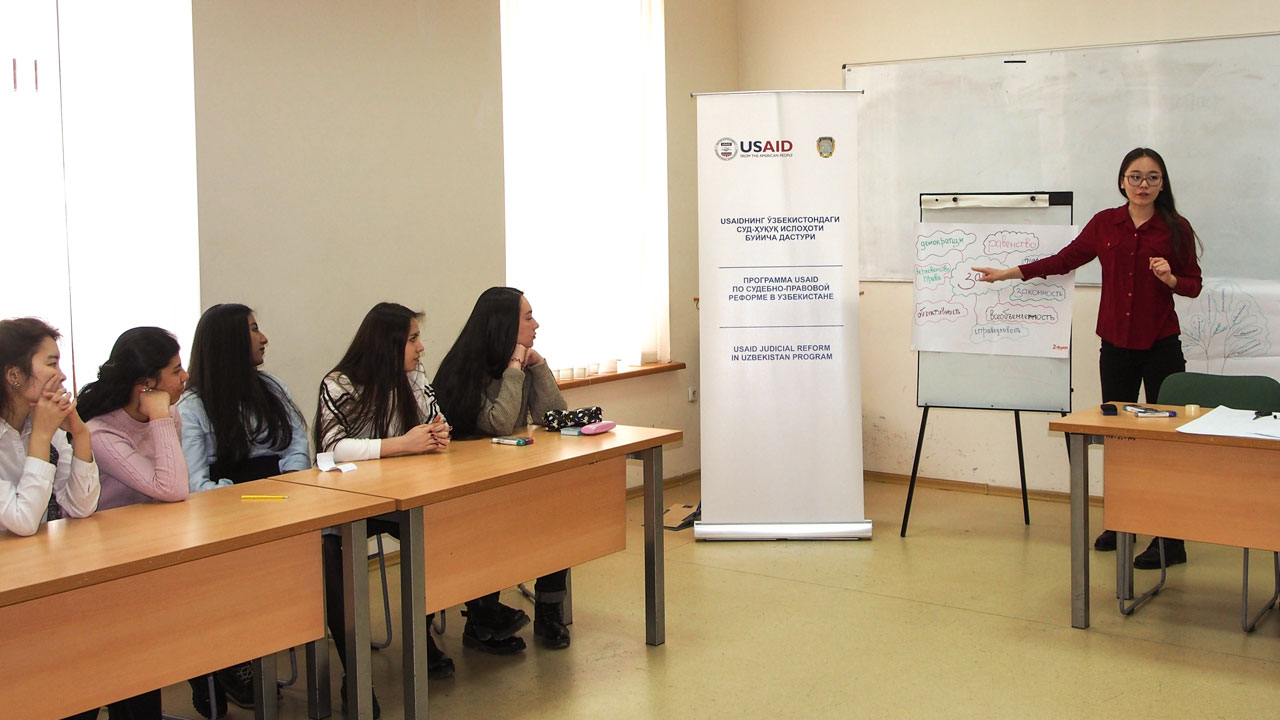 A lawyer, supported by the Tetra Tech-led Uzbekistan Justice and Rule of Law program, stands at the front of a meeting room presenting findings on a poster to a group of five people