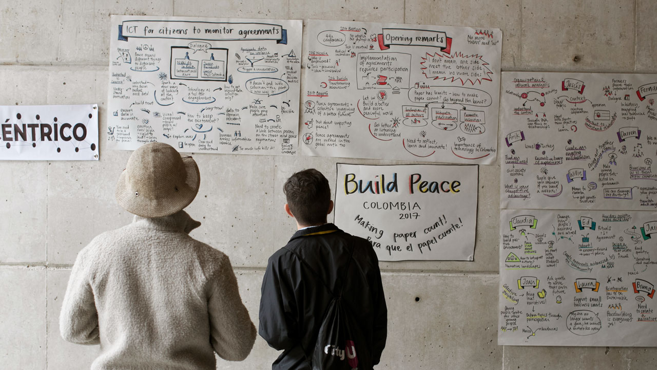 Two people stand in front of a wall with handwritten posters and graphics including one that reads “Build Peace Colombia” as part of Tetra Tech’s community peacebuilding activities