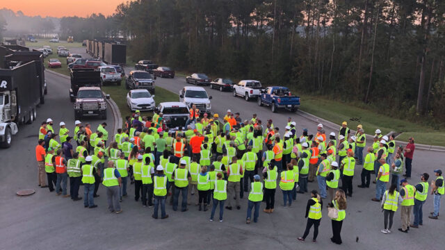 Group of field crew employees standing in road, wearing vests and hardhats, surrounded by work trucks