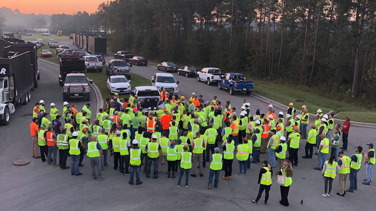 Group of field crew employees standing in road, wearing vests and hardhats, surrounded by work trucks