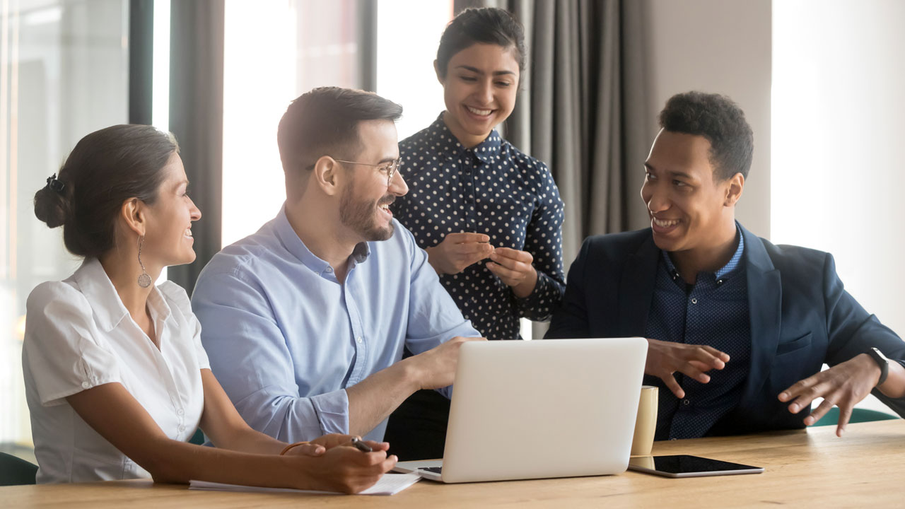 A diverse group of people sits around a conference table looking at a laptop and talking