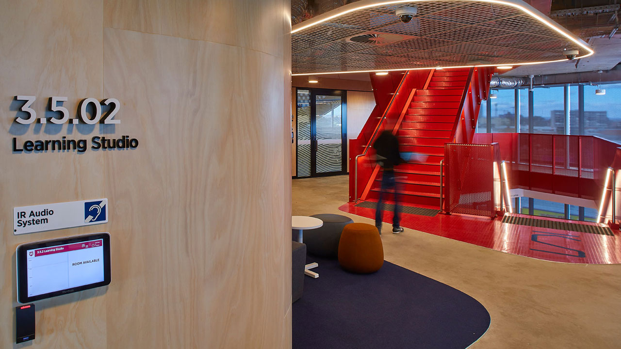 In the foreground is lettering on a wall that says "learning studio" with a student in the background walking up a staircase