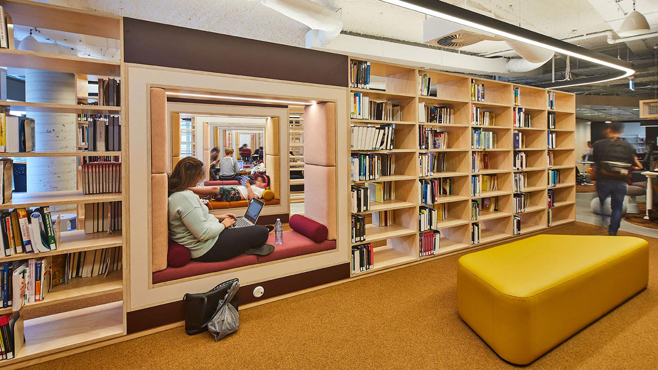 A university library with students working at their laptops beside a bookshelf
