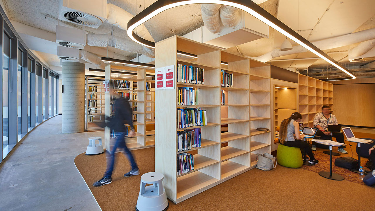 A university library. One student is walking from between the stacks in the middle ground. In the foreground students are seated with laptops