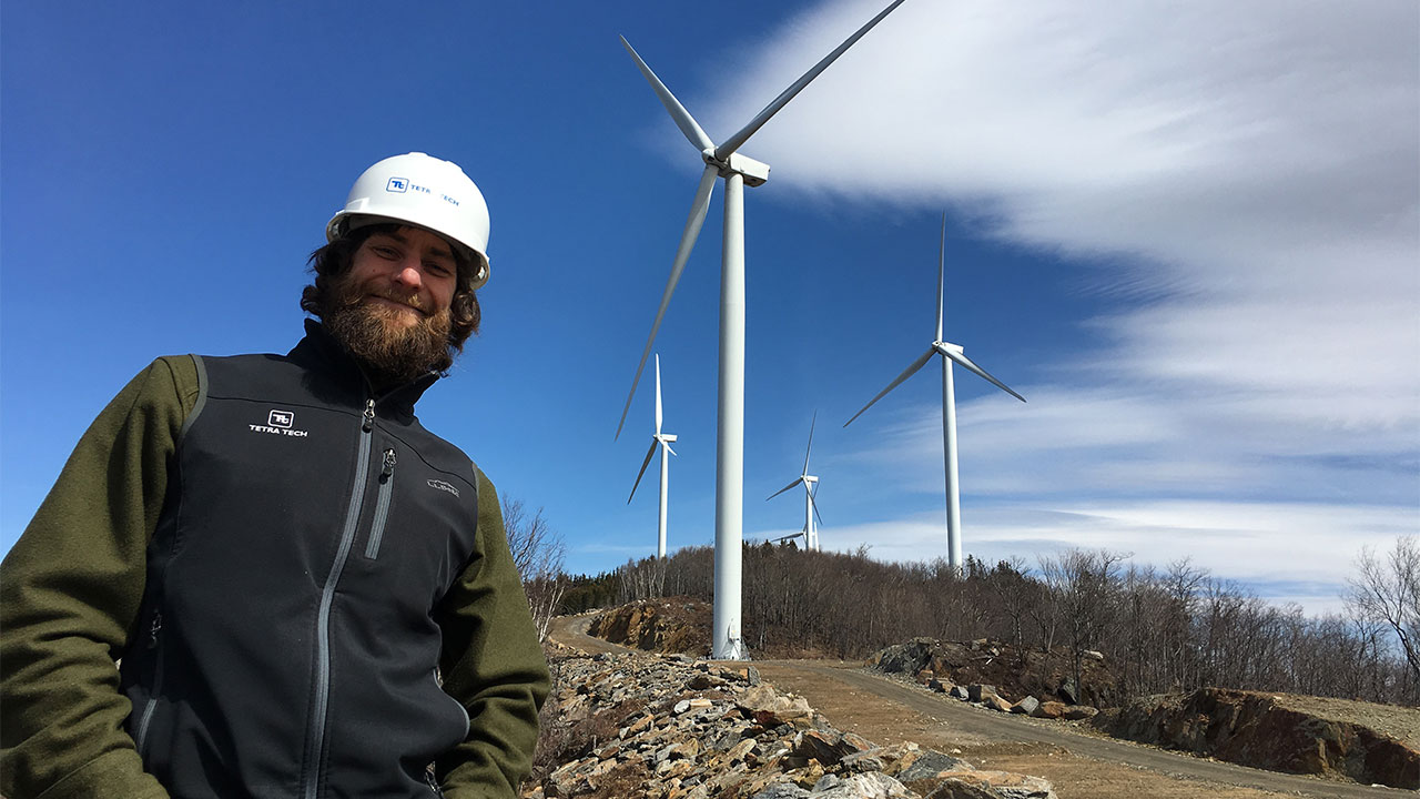 Tetra Tech employee in a white hard hat with four wind turbines in the background under a vast blue sky with white clouds