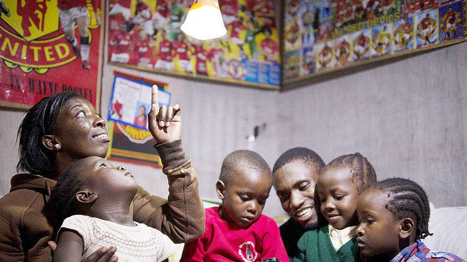 A family of six sits in their living room, newly lit from solar panels provided by the Tetra Tech-managed ACE program