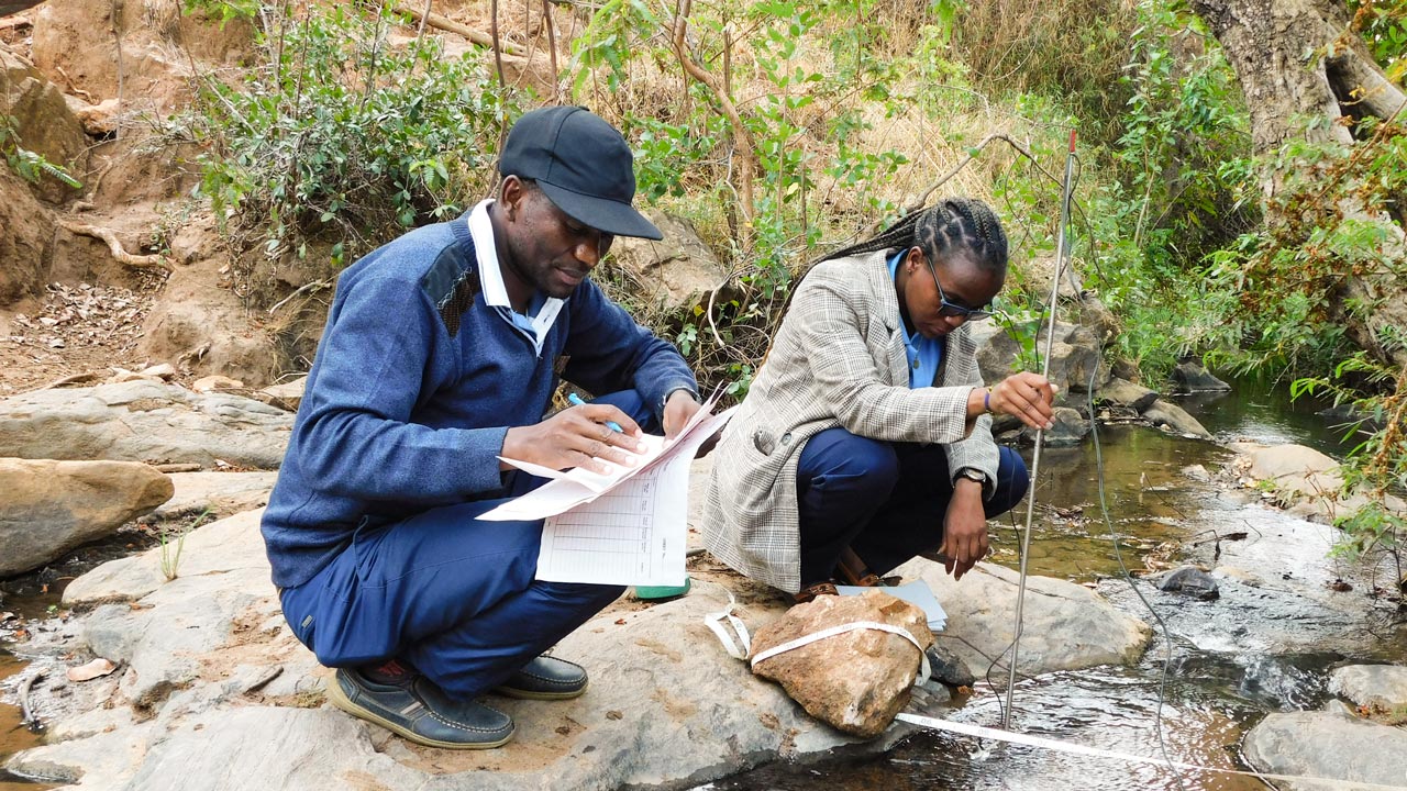 Two people sit on a rock beside a stream and measure the quality and quantity of the water