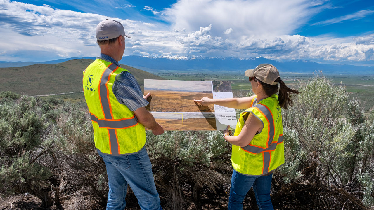 Two workers wearing hats and safety vests reviewing photo simulations in the field