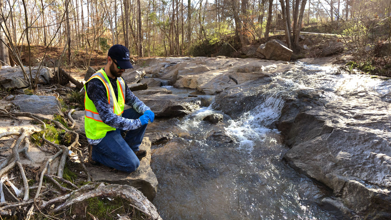 Worker wearing safety gear kneeling on a rock preparing to collect a stream water sample