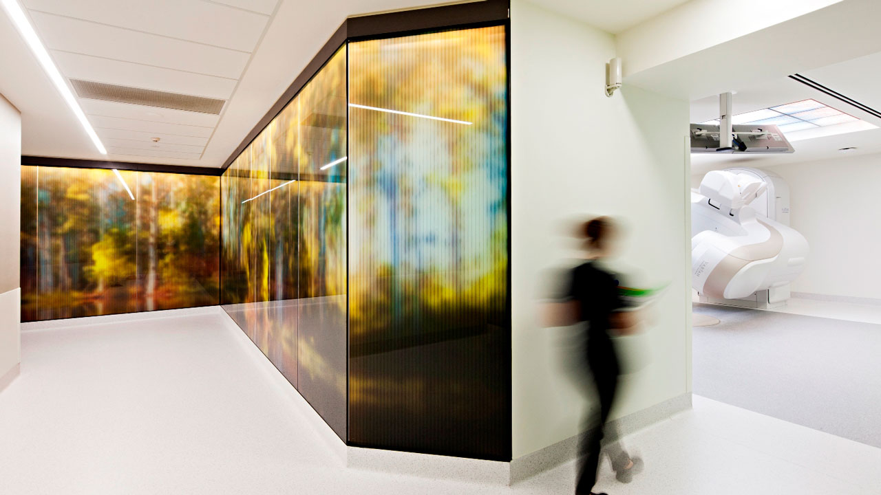 A medical professional walking up a corridor with a colored glass wall towards an open CT scanning room