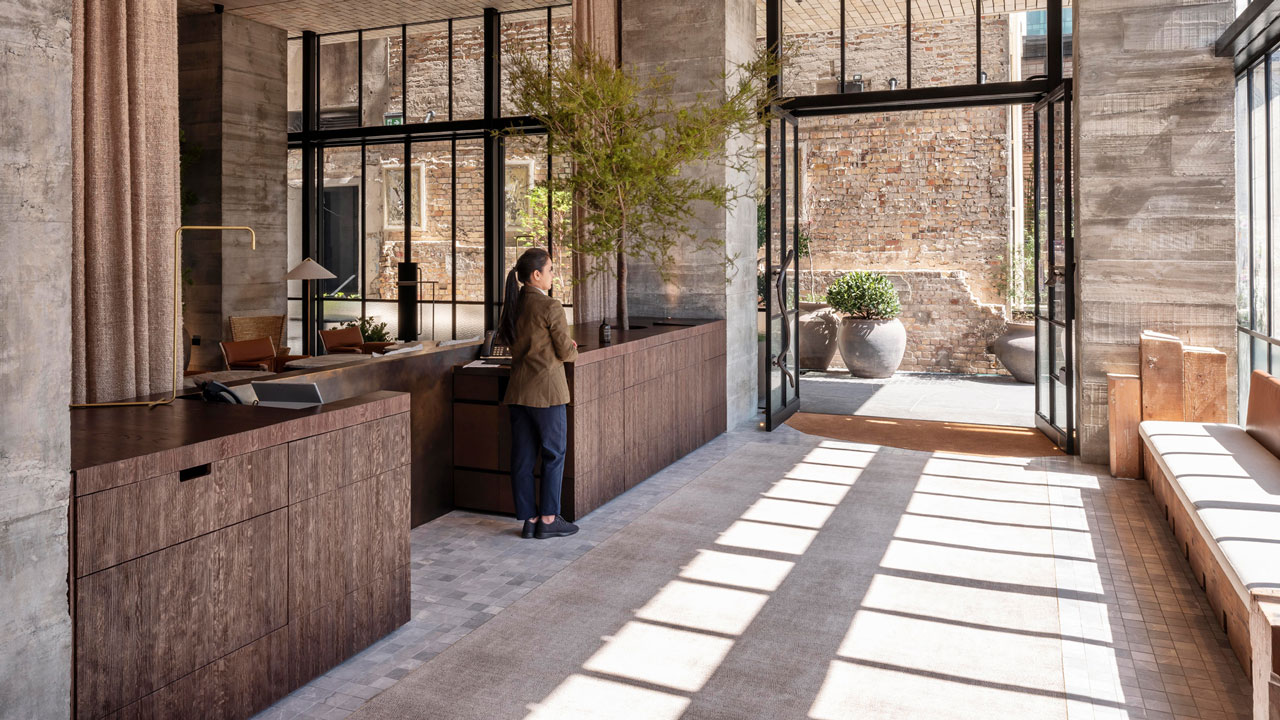 A hotel lobby with a bench seat under high windows, a woman standing at a long desk, and a walkway leading to large doors open to a terrace