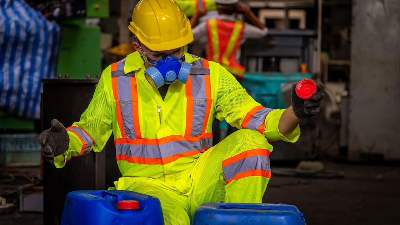 An engineer wearing safety uniform, black gloves, gas mask, and hard hat checking chemical tank in a factory