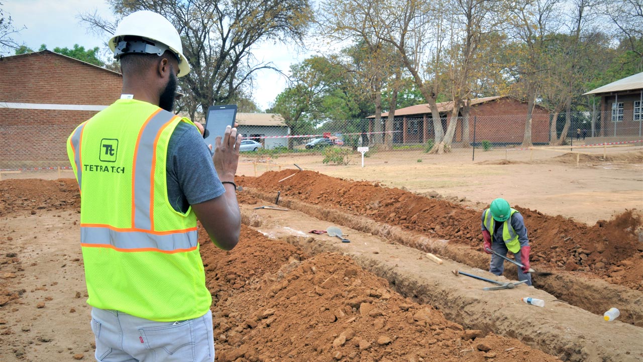 Man with a Tetra Tech high visibility vest holds a tablet and watches a man digging a trench in Malawi