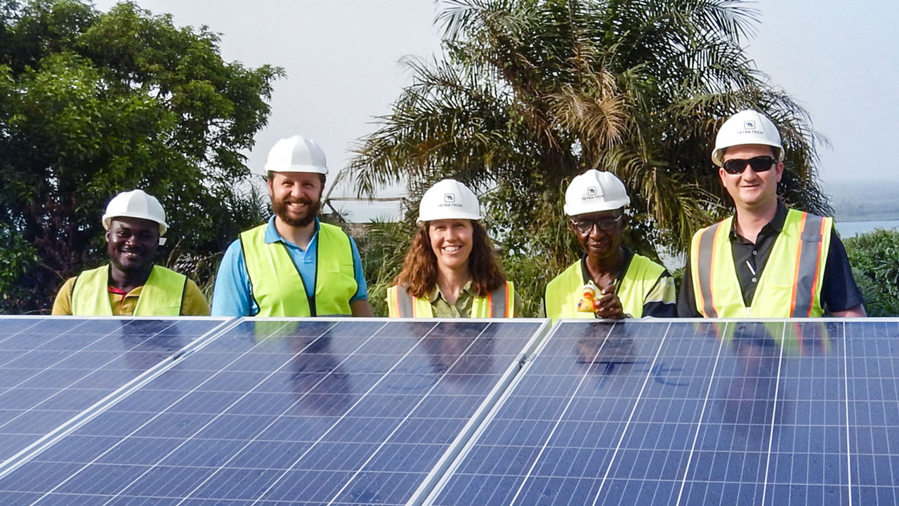 Five people in hard hats and high visibility vests stand behind a solar panel