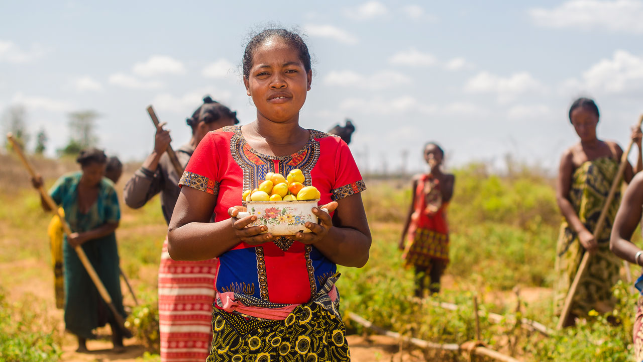 Woman holding a bowl of fruits with several people in the background farming a field