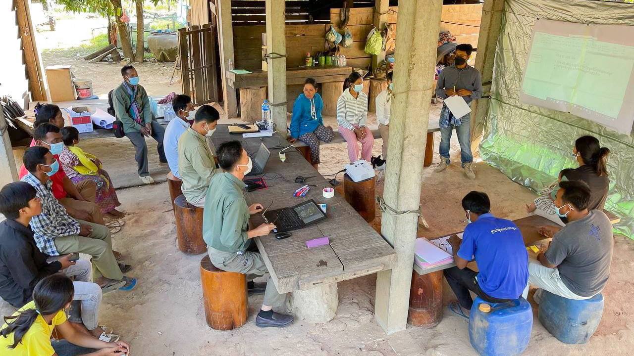A group of people in Cambodia sit underneath a structure at tables with laptops and notebooks
