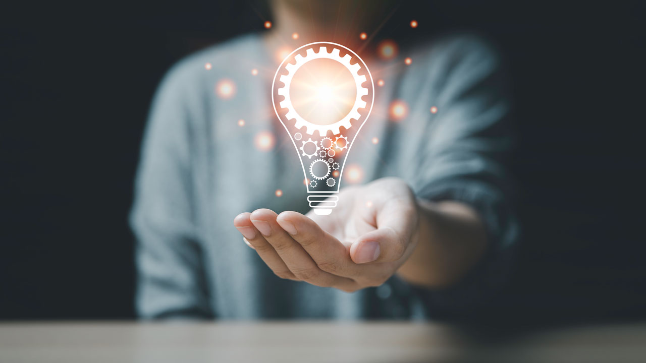 A digital rendering of a lightbulb with gears in glowing light gold floats above the outstretched, cupped hand of a person in a dark room