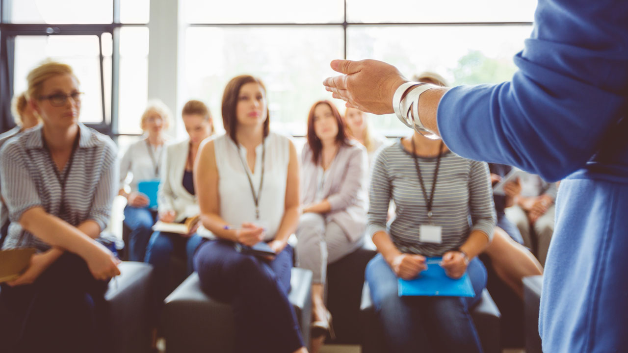 A blurred view of people sitting in a casual classroom, facing a presenter wearing a blue jacket whose arm and side are shown in the foreground