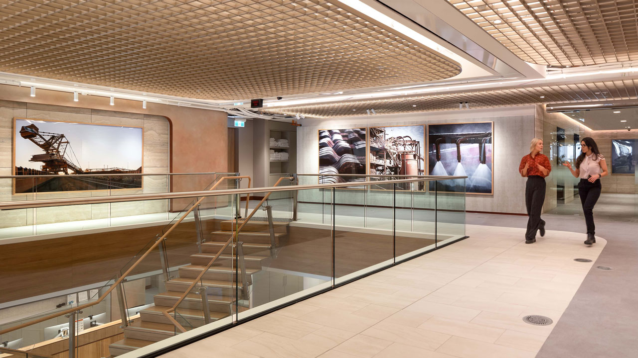 Two women walk through a spacious open area near a stairway with glass surround with wide lighting panels across the ceiling