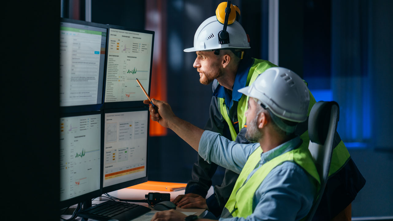 Two people in PPE in an industrial setting look at a bank of computer monitors