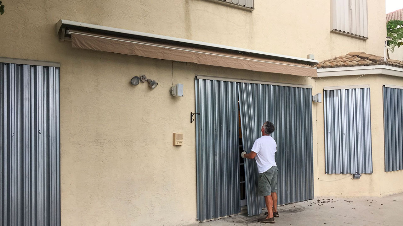 Man wearing gloves installing metal hurricane shutters on tan building