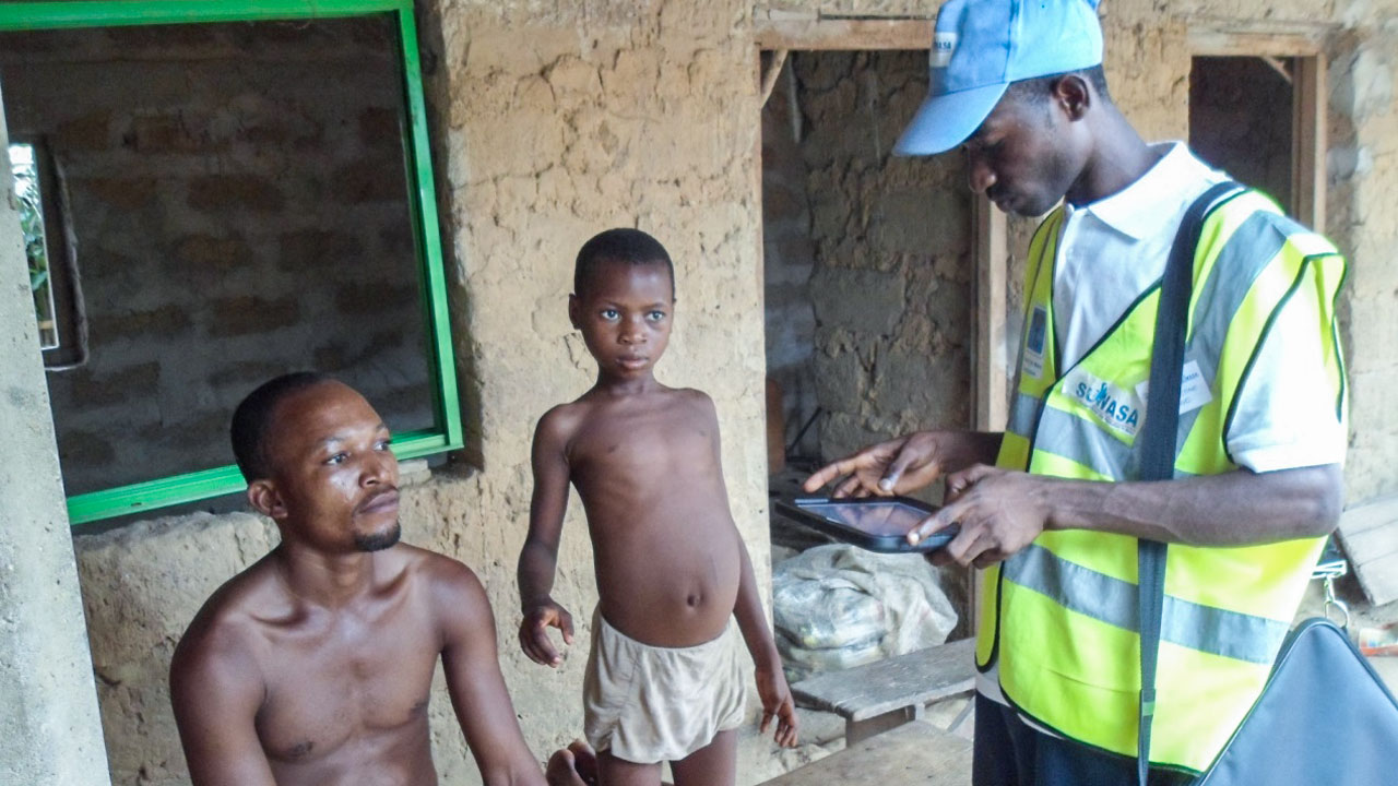 Man in a high visibility vest conducts a household survey, entering data on his tablet while a man and his son sit and watch
