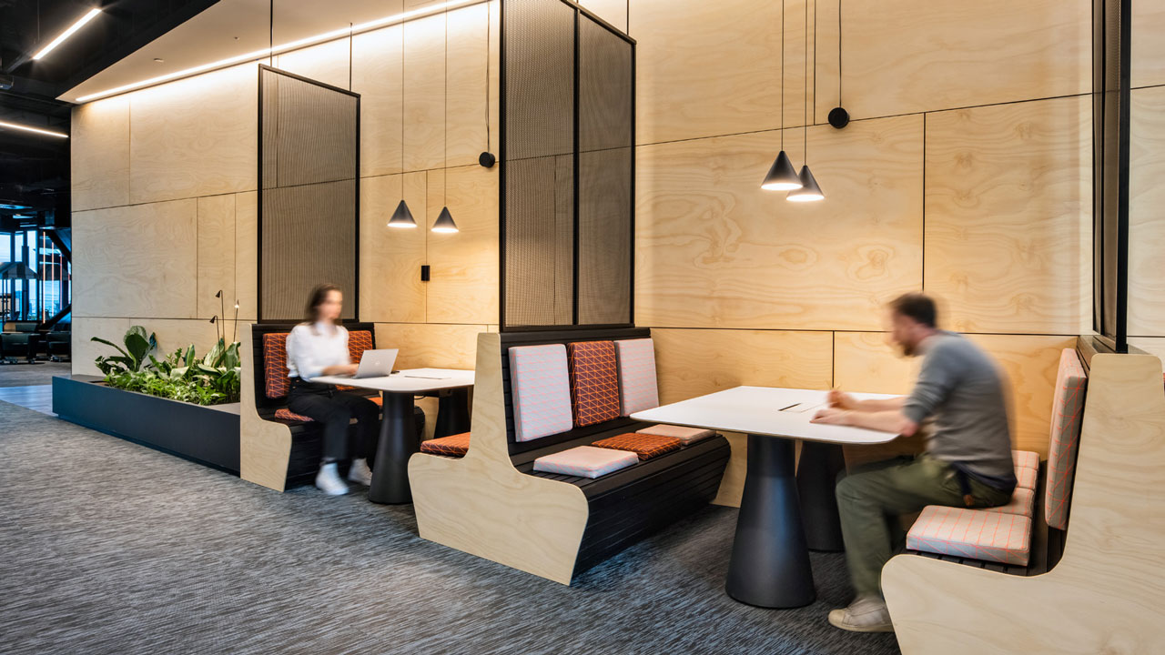 A modern workplace with people at booth tables with timber walls and feature lighting hanging from the ceiling