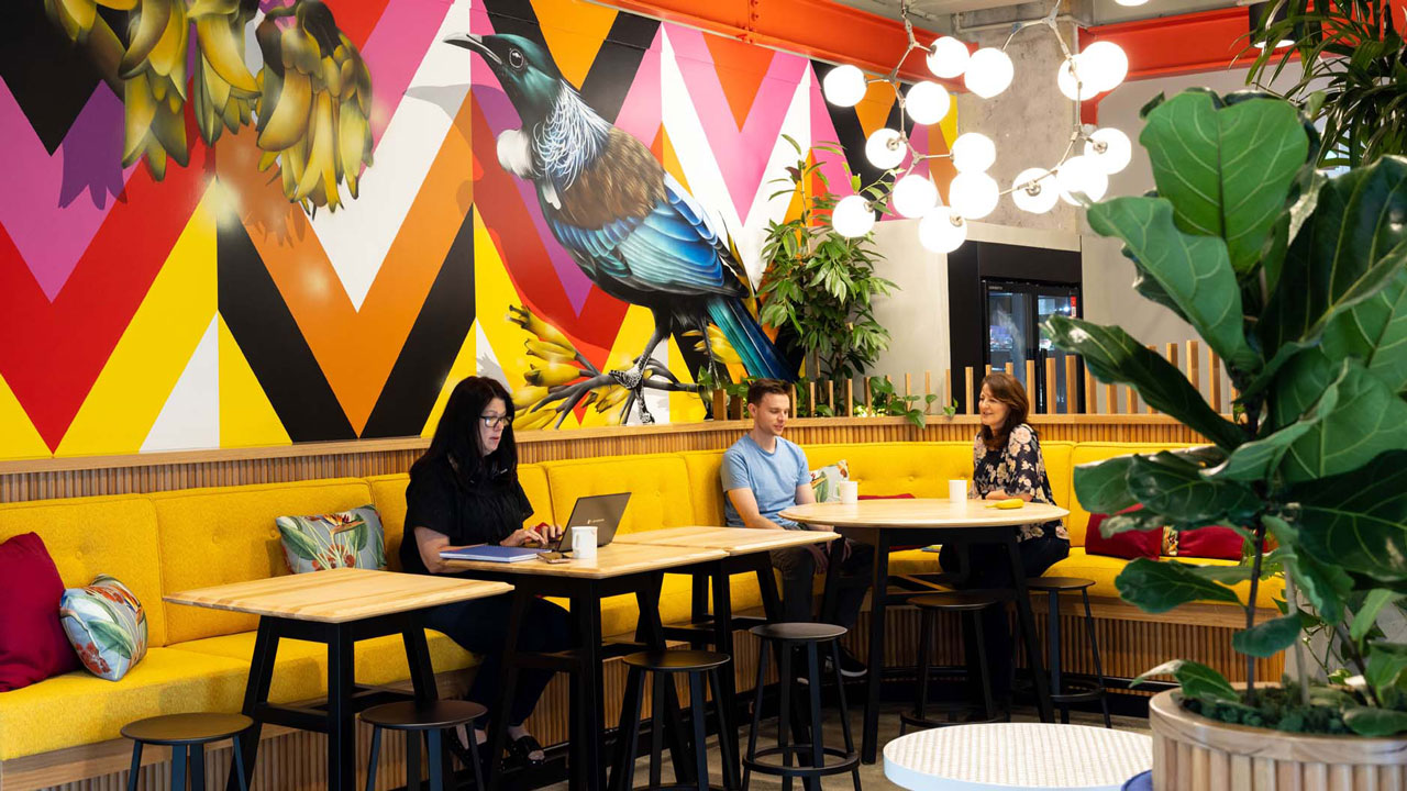 Three people at tables in a café-style workspace in an office with colorful artwork of birds and flowers on the wall behind them
