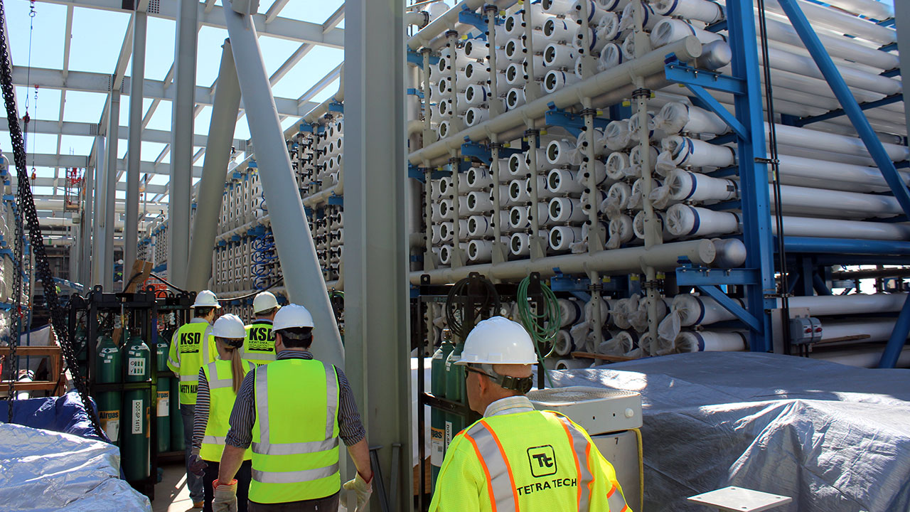 Tetra Tech employees wearing lime safety vests and hard hats walking in a line next to a stack of pipes