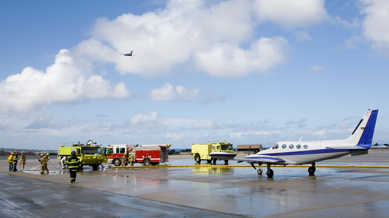 Fire trucks and firefighters on the tarmac with a small aircraft under a vast blue sky with fluffy clouds
